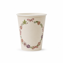 A cream-colored paper cup decorated with a delicate floral wreath pattern featuring pink, purple, and green elements, set against a plain white background.