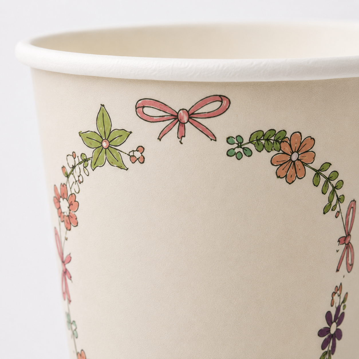 A close-up of a cream-colored paper cup decorated with a delicate border of pink bows, green leaves, and small orange and purple flowers.