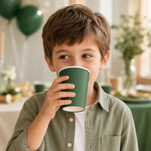 A young boy with brown hair wearing a sage green button-up shirt and white undershirt, holding a dark green paper cup to his mouth in a softly lit party setting with balloons in the background.