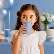 A young girl with long brown hair wearing a white ruffled dress holds a light blue paper cup to her mouth at a birthday party. The background features a blue table, balloons, flowers, and a decorated cake.