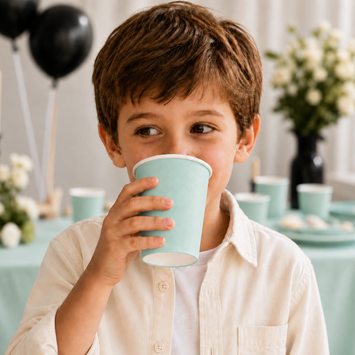 A young boy with brown hair wearing a cream-colored button-up shirt is holding a light blue paper cup to his lips. The background shows a softly blurred party setup with matching light blue cups, black balloons, and white flowers.