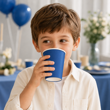 A young boy with brown hair wearing a cream-colored button-up shirt, holding a blue paper party cup to his mouth in a decorated party setting with blue balloons and a blue tablecloth in the background.