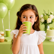 A young girl with long brown hair wearing a white ruffled dress holds a light green paper cup to her mouth. She is standing in a decorated party setting with a light green background, balloons, cupcakes, and a cake.