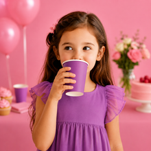A young girl with long brown hair wearing a purple dress is shown drinking from a matching purple paper cup in a pink-themed party setting with balloons, cupcakes, and a cake.