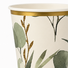 Close-up of a paper cup with a white interior, a metallic gold rim, and a botanical print featuring green leaves and gold glittery stems on an off-white background.