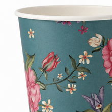A close-up of a paper cup with a teal background and a floral print featuring pink roses, white flowers, green leaves, and purple blossoms. The rim of the cup is white.